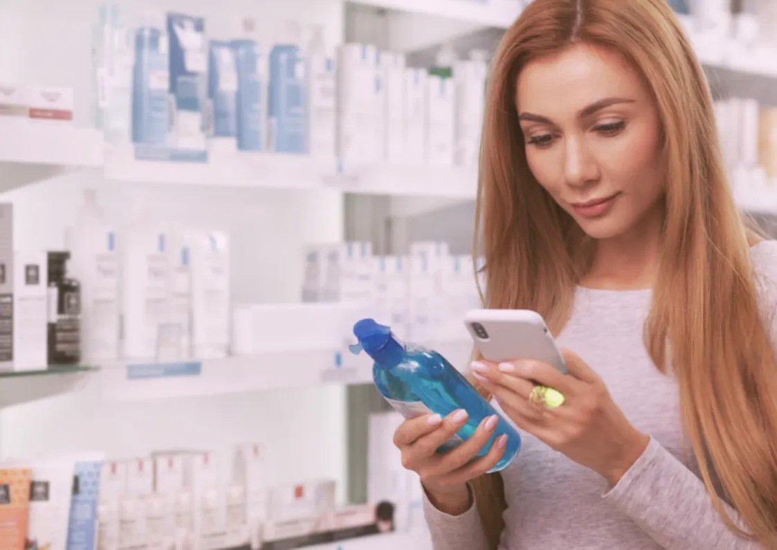 A woman in a pharmacy looks at a blue bottle while using her smartphone, with shelves of skincare products in the background.