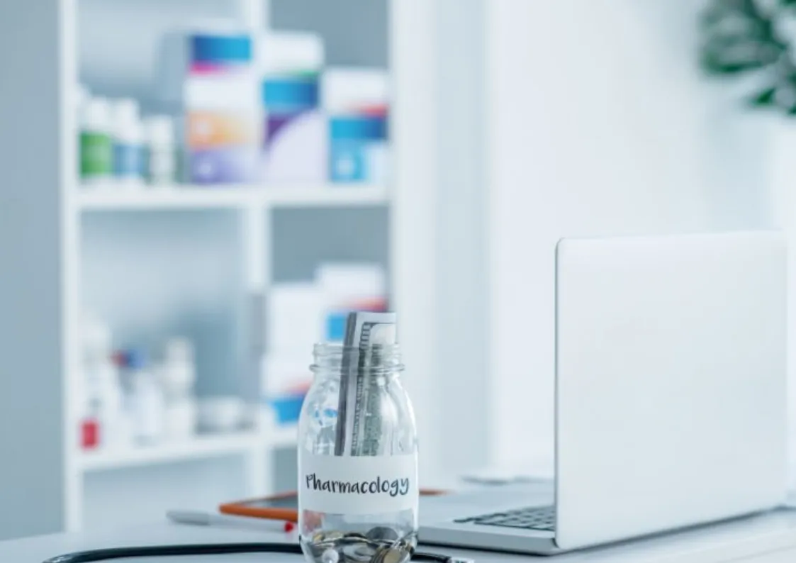 A laptop and a jar labeled "Pharmacology" are on a desk. Shelves with various bottles are blurred in the background.